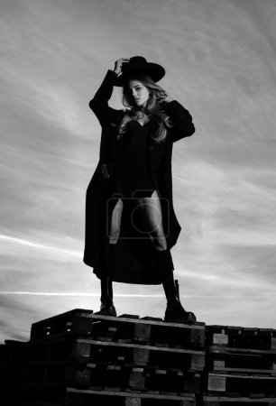 Téléchargez les photos : Femme sexy en manteau de mode noir, chapeau et chaussures noires bottes. Photo extérieure de mode de magnifique femme sensuelle dans des vêtements élégants. Beauté et fille de la mode - en image libre de droit