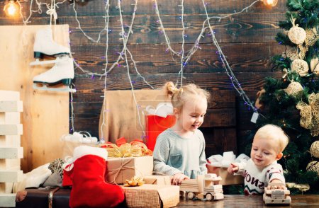 Téléchargez les photos : Joyeux petit enfant habillé en vêtements d'hiver pense au Père Noël près de l'arbre de Noël. Un enfant souriant qui regarde derrière un sapin de Noël. Joyeux enfant avec un cadeau de Noël sur fond en bois - en image libre de droit