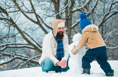 Téléchargez les photos : Heureux père et fils faisant bonhomme de neige dans la neige. Bonhomme de neige drôle fait main. Joyeux famille plaçant avec un bonhomme de neige sur une promenade hivernale enneigée - en image libre de droit
