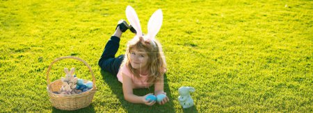 Téléchargez les photos : Enfants de Pâques garçon dans les oreilles de lapin chasse oeufs de Pâques en plein air. Enfant mignon en costume de lapin avec des oreilles de lapin s'amusant dans le parc. Cadre de bannière web panoramique - en image libre de droit
