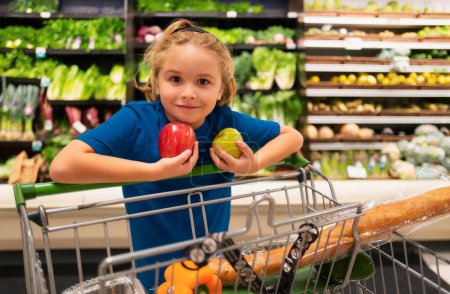 Téléchargez les photos : Enfant avec des pommes à l'épicerie. Faire du shopping avec des enfants. Un gamin qui achète des fruits au supermarché. Le petit garçon achète des légumes frais à l'épicerie. Un gamin qui choisit des légumes. Aliments sains - en image libre de droit