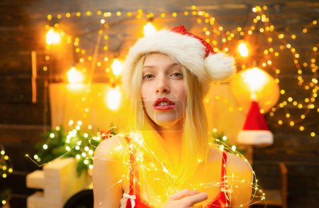Photo for Woman with red lips in christmas light is looking at camera. Girl is wearing Santa hat. Concept of holidays. Christmas mood - Royalty Free Image