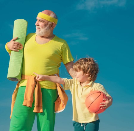 Foto de Feliz familia amorosa. Sportsman abuelo y niño sano con pelota de baloncesto sobre fondo azul cielo. Deporte para niños pequeños - Imagen libre de derechos