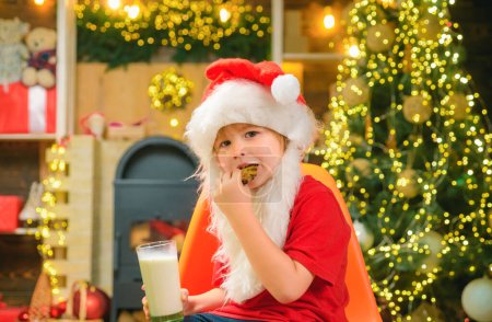Téléchargez les photos : Père Noël enfant manger des biscuits et boire du lait. Père Noël garçon en chapeau de Père Noël - en image libre de droit