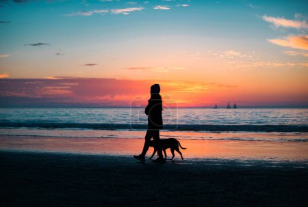 Foto de Mujer paseando con el perro en la playa. Hermoso amanecer, sol, cielo azul con nubes y el paisaje marino - Imagen libre de derechos
