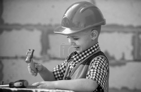 Téléchargez les photos : Enfant en casque dur tenant le marteau. Petit enfant aidant avec des outils de jouet sur le site construciton. Des enfants avec des outils de construction. Travailleur de la construction. Réparation maison - en image libre de droit