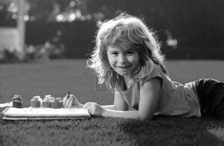 Téléchargez les photos : Dessin d'enfant. Kid dessine dans le parc posé dans l'herbe en s'amusant sur fond de nature - en image libre de droit