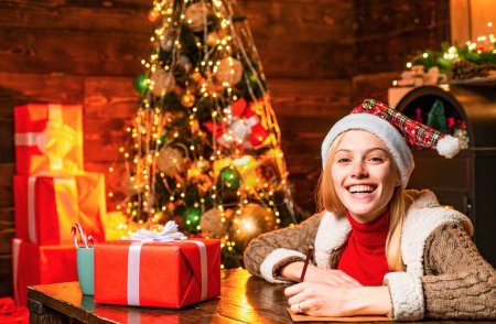 Photo for Cheerful smiling blonde woman writing a letter to Santa with her wish list of presents. Christmas presents gifts concept. Joy and happiness. Christmas. Xmas tree - Royalty Free Image