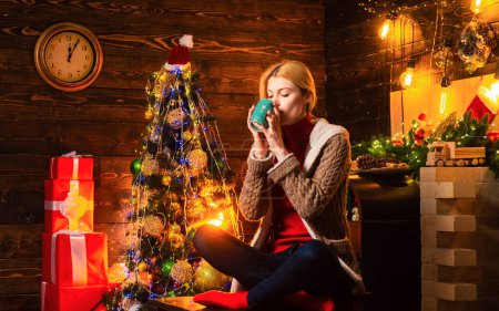 Foto de La señora está bebiendo bebida caliente y sentada junto al árbol de Navidad. Feliz jovencita junto a la chimenea. Invierno, Navidad - Imagen libre de derechos
