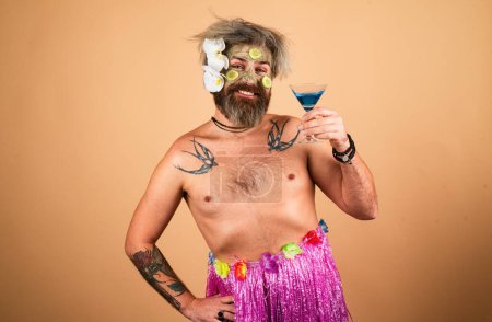 Téléchargez les photos : Homme drôle à Hawaï. Modèle masculin avec masque en argile et tranches de concombre sur le visage. Homme ayant masque hydratant cosmétique fleurs hawaïennes boire cocktail, fond studio isolé. Soins du visage masculin - en image libre de droit