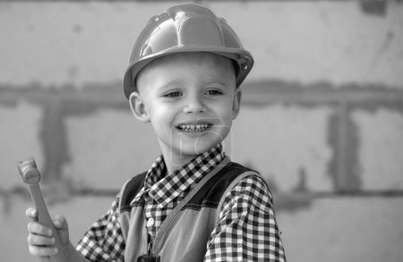Foto de Un chico con sombrero duro sosteniendo el martillo. Feliz niño sonriente ayudando con herramientas de juguete en el sitio de construciton. Niños con herramientas de construcción. Trabajadores de la construcción - Imagen libre de derechos