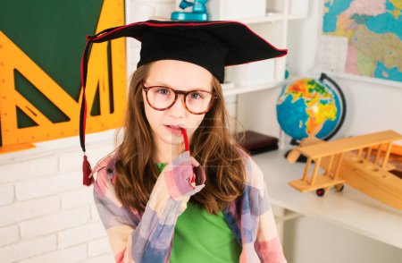 Photo for Happy girl with pen in mouth in graduation cap near a school board - Royalty Free Image