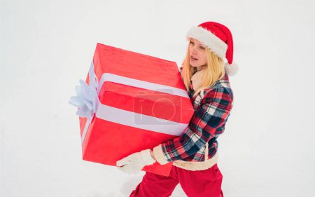 Téléchargez les photos : Femme heureuse en robe rouge s'amusant avec une grande boîte cadeau isolée sur un fond de neige blanche. Cadeau de Noël emballé avec ruban. Bonne période hivernale - en image libre de droit
