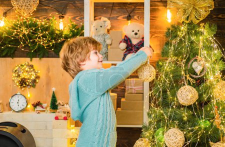 Foto de Niño en invierno vestido de Navidad decorando el árbol de Navidad con adornos. Niño decoración árbol de Navidad y divertirse - Imagen libre de derechos