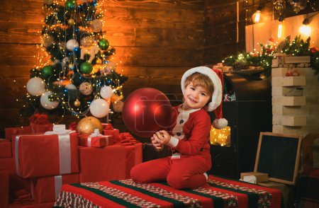 Téléchargez les photos : Un enfant heureux porte des vêtements de père Noël tenant une grosse boule rouge. Concept enfant de Noël. Vacances - en image libre de droit