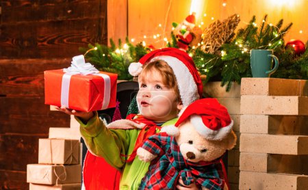 Foto de Niño Santa sosteniendo y mirando regalo de la caja de Navidad tratando de adivinar lo que hay dentro de ella. Un niño en el sombrero de Santas abrazó a su bonito osito de peluche. El niño de Navidad sueña. Dulce hogar - Imagen libre de derechos