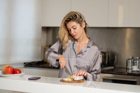Téléchargez les photos : Sexy femme au foyer prendre le petit déjeuner à la cuisine. Style de vie domestique. Femme sensuelle coupant fromage et saucisse dans la cuisine le matin - en image libre de droit