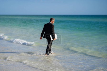 Téléchargez les photos : Homme d'affaires en costume avec ordinateur portable dans l'eau de mer. Tourisme de voyage et concept d'affaires. Employé de bureau fou avec ordinateur portable sur la plage. Vue arrière du dos homme d'affaires en costume dans l'eau de mer à la plage - en image libre de droit