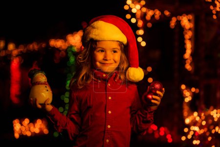 Téléchargez les photos : Enfant debout près de la maison illuminée de Noël de nuit. Enfant en chapeau de Père Noël près des guirlandes de la maison dehors. Maison décorée avec des guirlandes - en image libre de droit