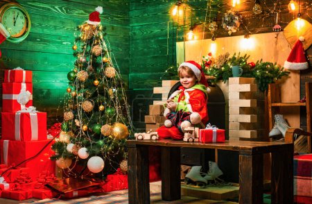 Photo for New Year. Little boy in Santa hat and costume having fun. Holiday discounts. Little boy decorating xmas tree and opening presents. Xmas tree. Child cheerful face got gift in christmas sock - Royalty Free Image