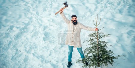 Foto de Alegre leñador osos abeto en el fondo de nieve blanca. Hombre divertido de Santa posando con hacha y árbol de Navidad. Joven leñador está cortando el árbol de Navidad en la madera - Imagen libre de derechos