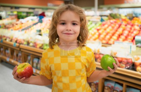 Téléchargez les photos : Un enfant tient des pommes à l'épicerie. Portrait d'enfant dans un magasin d'alimentation ou un supermarché. Un gamin qui va faire du shopping. Aliments sains pour enfants - en image libre de droit