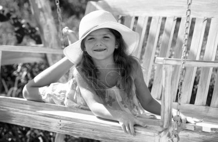 Téléchargez les photos : Joyeux enfant fille riant et se balançant sur une balançoire au jardin d'été. Petite fille qui s'amuse sur une balançoire extérieure. Portrait d'une belle enfant fille en chapeau rêvant - en image libre de droit