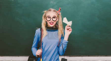 Photo for Portrait of a teen female student. School teenager girl holding backpack and paper plane, standing against blackboard - Royalty Free Image