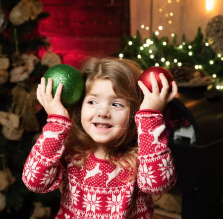 Photo for Little girl in red jacket with snowflakes and deers. Holiday concept. Happy little girl preschooler in a winter sweater is holding red and green Christmas tree balls. Christmas time - Royalty Free Image