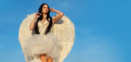 Téléchargez les photos : Sensuelle fille en costume d'ange robe blanche et ailes de plumes sur fond de ciel. En-tête de bannière avec espace de copie - en image libre de droit
