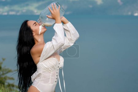 Foto de Mujer sexy en ropa interior beber leche de lata y botella contra el campo. Mujer sexy bebiendo leche en los Alpes. Mujer sensual comer leche cerca de los Alpes suizos. Hermosa mujer disfruta de la leche en el pueblo alpino - Imagen libre de derechos