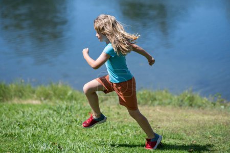 Foto de Niño corriendo sobre hierba verde cerca del lago en el parque de verano. Niño corriendo al aire libre. Actividad deportiva saludable para niños. Niño en la carrera de atletismo. Corredor niños haciendo ejercicio - Imagen libre de derechos