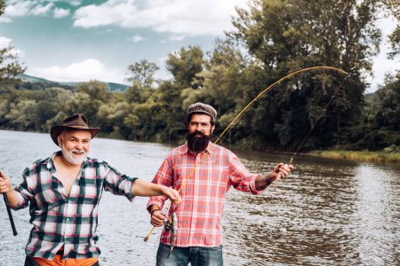 Foto de Dos hombres amigos pescando. Pesca con mosca pescador hace yeso, de pie en el agua del río. Viejo y joven pescador - Imagen libre de derechos