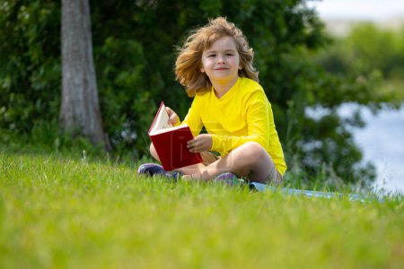 Foto de Kid read a book. Early education for kids. Summer vacation with reading book. Outdoor homework. Summer camp with book. Kids reading and education concept. Kid sitting on grass and reading a book - Imagen libre de derechos