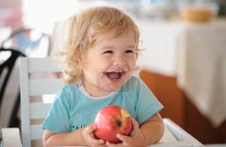 Téléchargez les photos : Rire mignon enfant mangeant de la pomme. Mignon bébé manger des pommes. Portrait de mignon sourire enfant caucasien assis dans la chaise haute manger des fruits de pomme - en image libre de droit