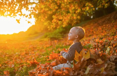 Foto de Smilimg linda niña sentada en las hojas cubiertas de la tierra viendo caer las hojas. Feliz infancia. Los primeros recuerdos. Volver a la seguridad, el cuidado y la tranquilidad - Imagen libre de derechos