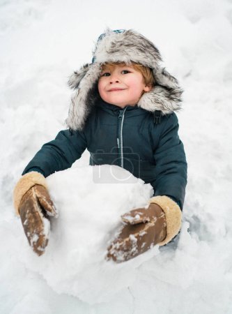Foto de Muñeco de nieve y niño divertido el amigo está de pie en sombrero de invierno y bufanda con nariz roja. Chico alegre divirtiéndose con muñeco de nieve en Winter Park. Ropa de invierno para niños - Imagen libre de derechos