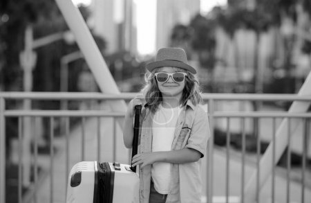 Foto de Divertido viajero infantil. Niño turista viajero en ropa casual, gafas de sol de moda y sombrero sostienen maleta. Viaja al extranjero los fines de semana. Viaje de niños - Imagen libre de derechos