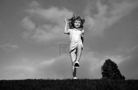 Téléchargez les photos : Petit garçon courant et sautant sur l'herbe près d'une prairie verte printanière sur ciel bleu. Fille enfant heureux jouant sur un terrain ensoleillé, été nature style de vie en plein air - en image libre de droit