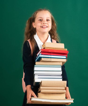 Photo for Smiling funny little schoolkid girl with backpack hold books on green blackboard. Childhood lifestyle concept. Education in school. Knowledge day - Royalty Free Image
