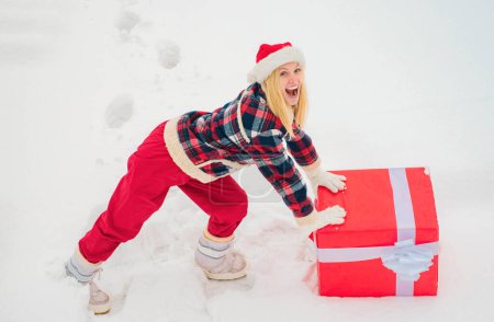 Foto de Mujer divertida de Navidad y una gran caja de regalo. Una mujer divertida sosteniendo un gran regalo de Navidad. Loca chica de Navidad empuja un gran regalo en la nieve fondo de invierno - Imagen libre de derechos