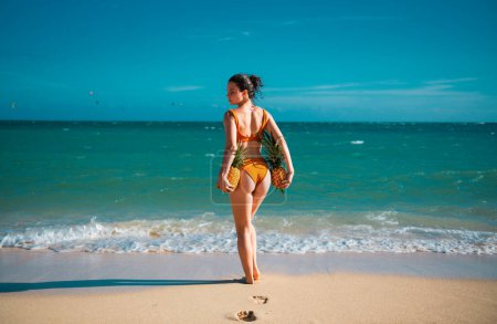 Téléchargez les photos : Femme avec des fesses sexy en maillot de bain. Jeune femme tenant un ananas. Vacances d'été chaudes sur la plage de jamaica ou miami - en image libre de droit