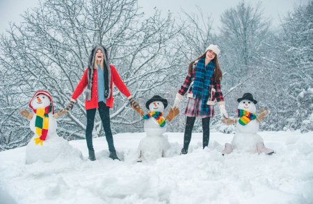Téléchargez les photos : Groupe de Beauté Winter Girls avec bonhomme de neige. Fête des étudiants et célébration de Noël. Deux joyeux beauté jeunes femmes s'amuser avec bonhomme de neige dans Winter Park. Joyeux Noël et bonne année - en image libre de droit