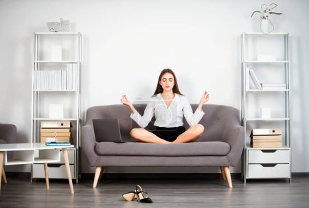 Foto de Joven mujer de negocios de meditación, secretaria haciendo ejercicio de yoga en el sofá en el lugar de trabajo en una oficina moderna. Empleado siente equilibrio armonía relajación - Imagen libre de derechos