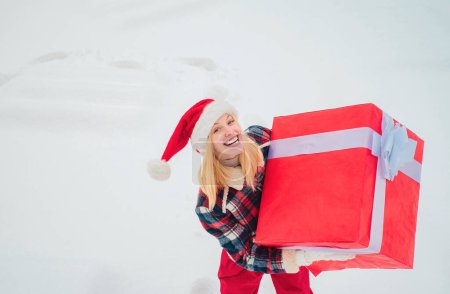 Téléchargez les photos : Service de livraison d'hiver. Femme portant un grand cadeau isolé sur fond d'hiver de neige. Portrait de Santa femme avec un énorme cadeau rouge regardant la caméra - en image libre de droit