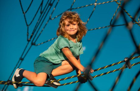 Foto de Lindo niño sonriente escalando la red en el patio de recreo. Parque de cuerdas para niños. Cara divertida de los niños - Imagen libre de derechos