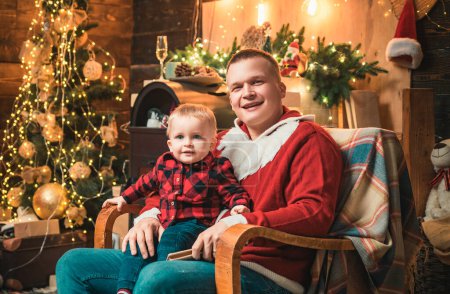 Téléchargez les photos : Portrait rapproché du père et du petit garçon heureux assis sur une chaise sur un fond en bois et un sapin de Noël dans la chambre décorée de luxe. Vacances d'hiver en famille et concept de personnes - en image libre de droit