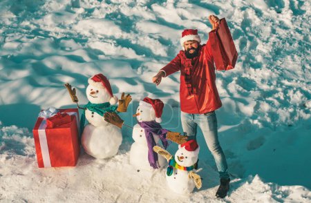 Téléchargez les photos : Beau Winter Man avec cadeau et bonhomme de neige dans le parc hivernal givré. Funny Santa homme posant avec boîte cadeau rouge sur le temps d'hiver. Vacances d'hiver et concept de personnes - en image libre de droit