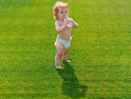 Foto de Bebé beber agua al aire libre. Niño pequeño caminando descalzo sobre hierba verde en la soleada noche de verano. Niño sano - Imagen libre de derechos