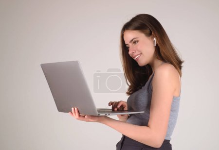 Téléchargez les photos : Joyeux élève. Femme Étudiante en t-shirt blanc tenant ordinateur portable et ordinateur portable, isolé sur fond gris. Étudiant posant en studio. Portrait d'un élève ayant un appel vidéo. Temps des étudiants - en image libre de droit